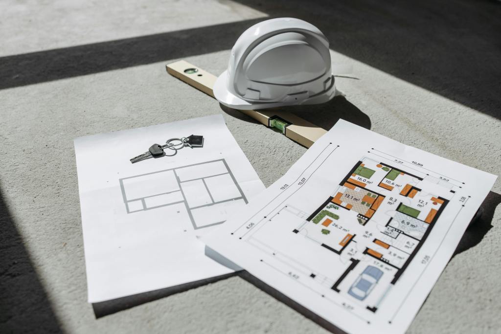 House floor plans laid on the ground with keys, a white hard hat, and a measuring level, representing construction and new housing development.