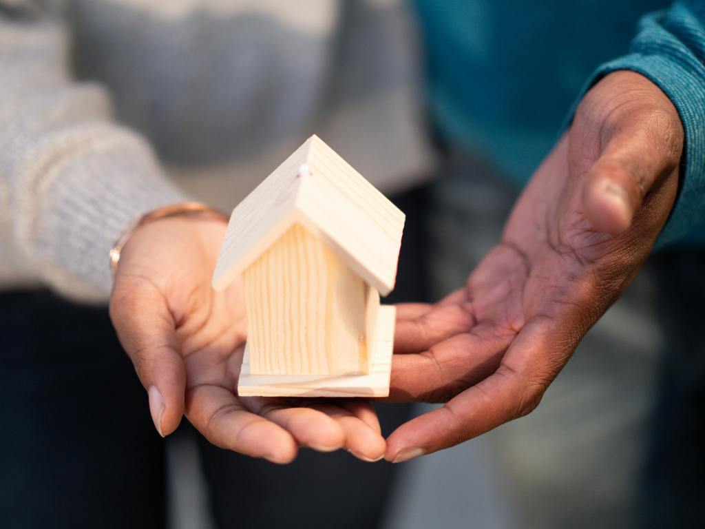 Close-up of two people holding a small wooden house together, symbolizing shared support and community.